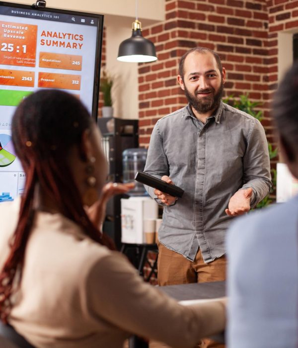 Executive manager standing beside monitor showing marketing graphs explaining company strategy during business meeting in startup office. Diverse businesspeople working at financial presentation