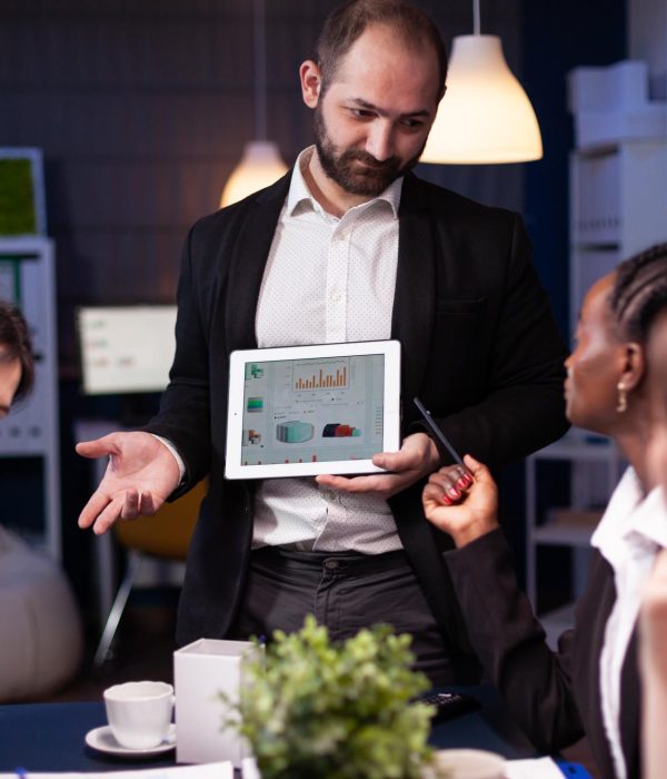 Focused workaholic entrepreneur man working overtime presenting company statistics using tablet. Diverse multi-ethnic businesspeople overworking in office meeting room late at night.